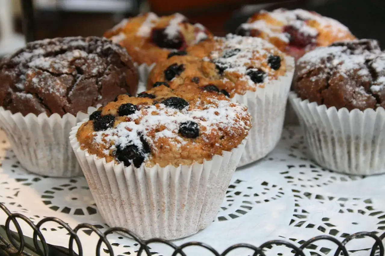 Flera vanliga muffins, n&aring;gra med bl&aring;b&auml;r och pudrade med florsocker, ligger p&aring; ett vitt spetsfat.