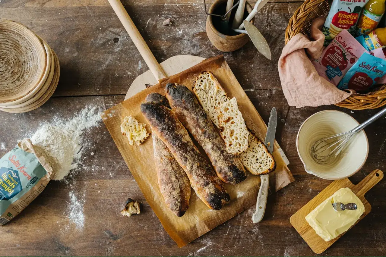 Nybakade surdegsbaguetter på bakplåtspapper, redo för ett surdegslimpa recept. Mjöl, smör och ingredienser för bakning ligger utspridda.