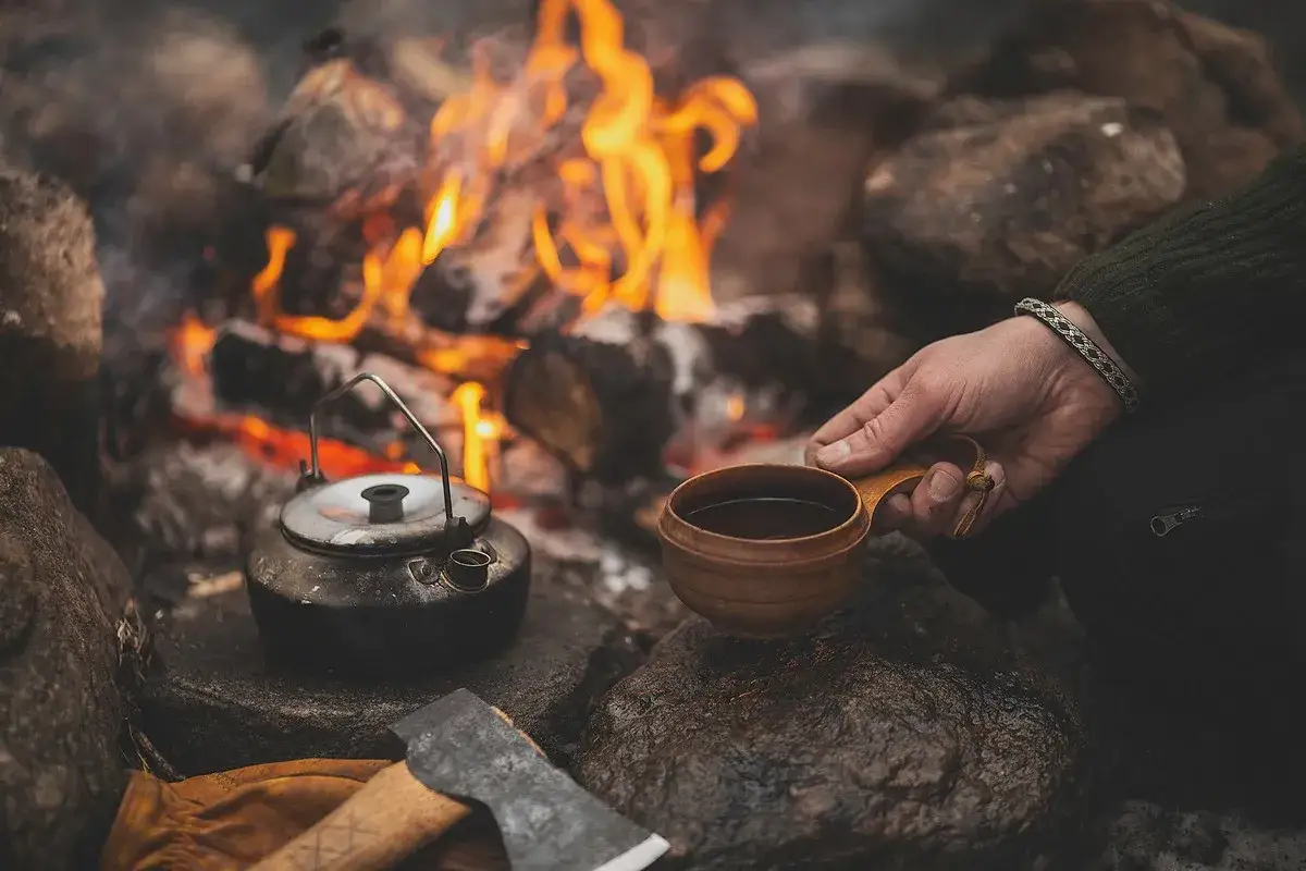 En hand håller en träkopp med kaffe vid en lägereld. Bredvid står en kaffepanna. Perfekt kokkaffe dosering för en mysig stund.
