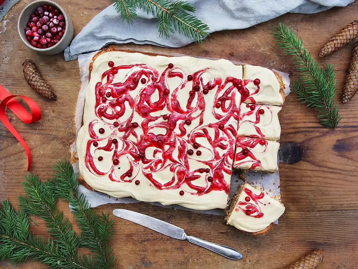 En skiva av mjuka pepparkakor med vit glasyr och lingon virvlar. Juliga dekorationer som granris och kottar omger bakverket.