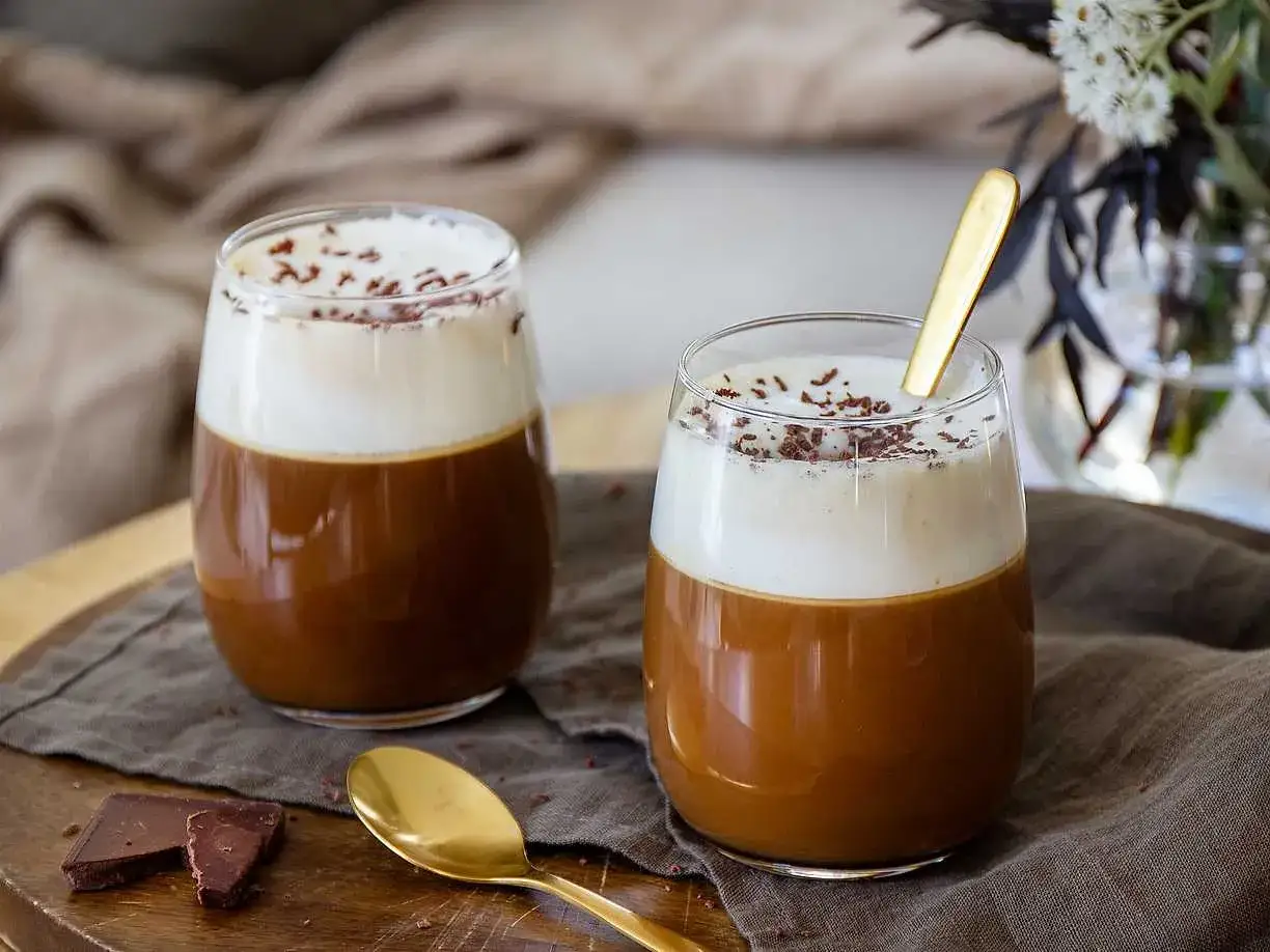 Tv&aring; glas med en kr&auml;mig kaffedrink, toppad med vispad gr&auml;dde och chokladstr&ouml;ssel. En gyllene sked vilar i det ena glaset, perfekt f&ouml;r en Leonard Sachs drink.