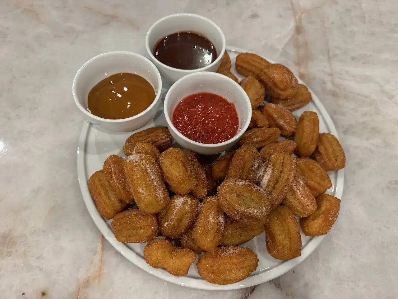 En tallrik med sockerströsslade churros, serverade med tre dippsåser: choklad, kolasås och jordgubbssylt. Perfekt för att njuta av vad är churros!