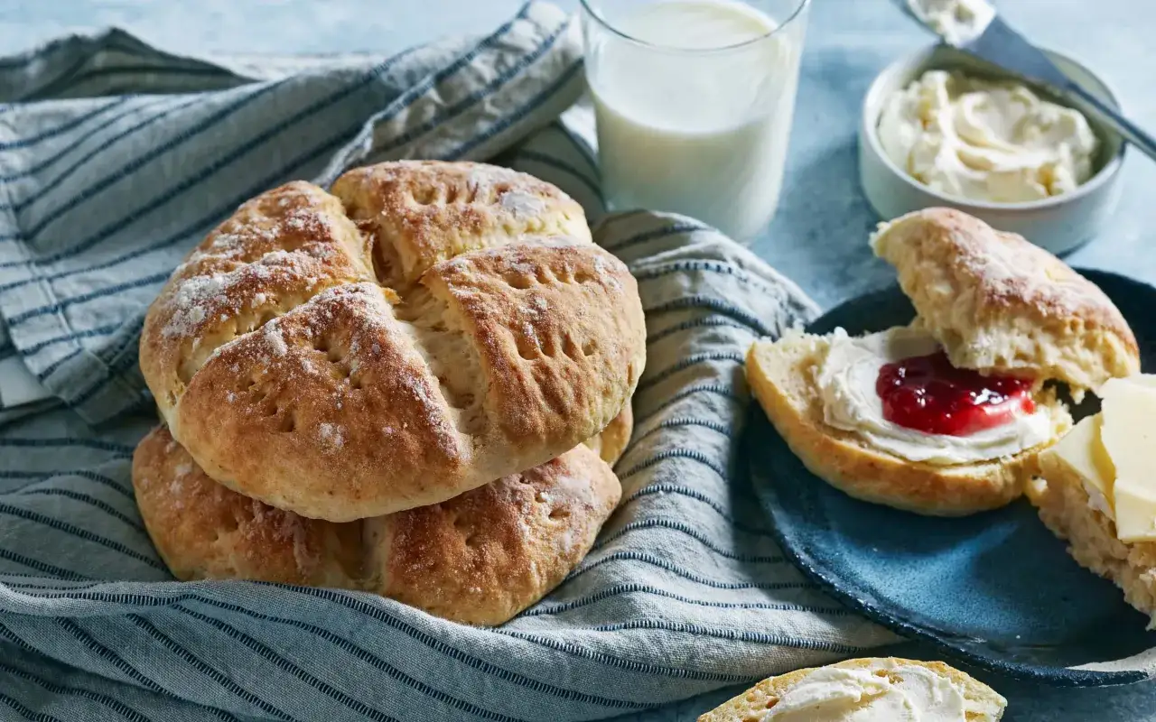 Nybakade engelska scones ligger på en randig duk. En halv scone är toppad med grädde och sylt, en annan med ost. Ett glas mjölk och en skål grädde finns i närheten.
