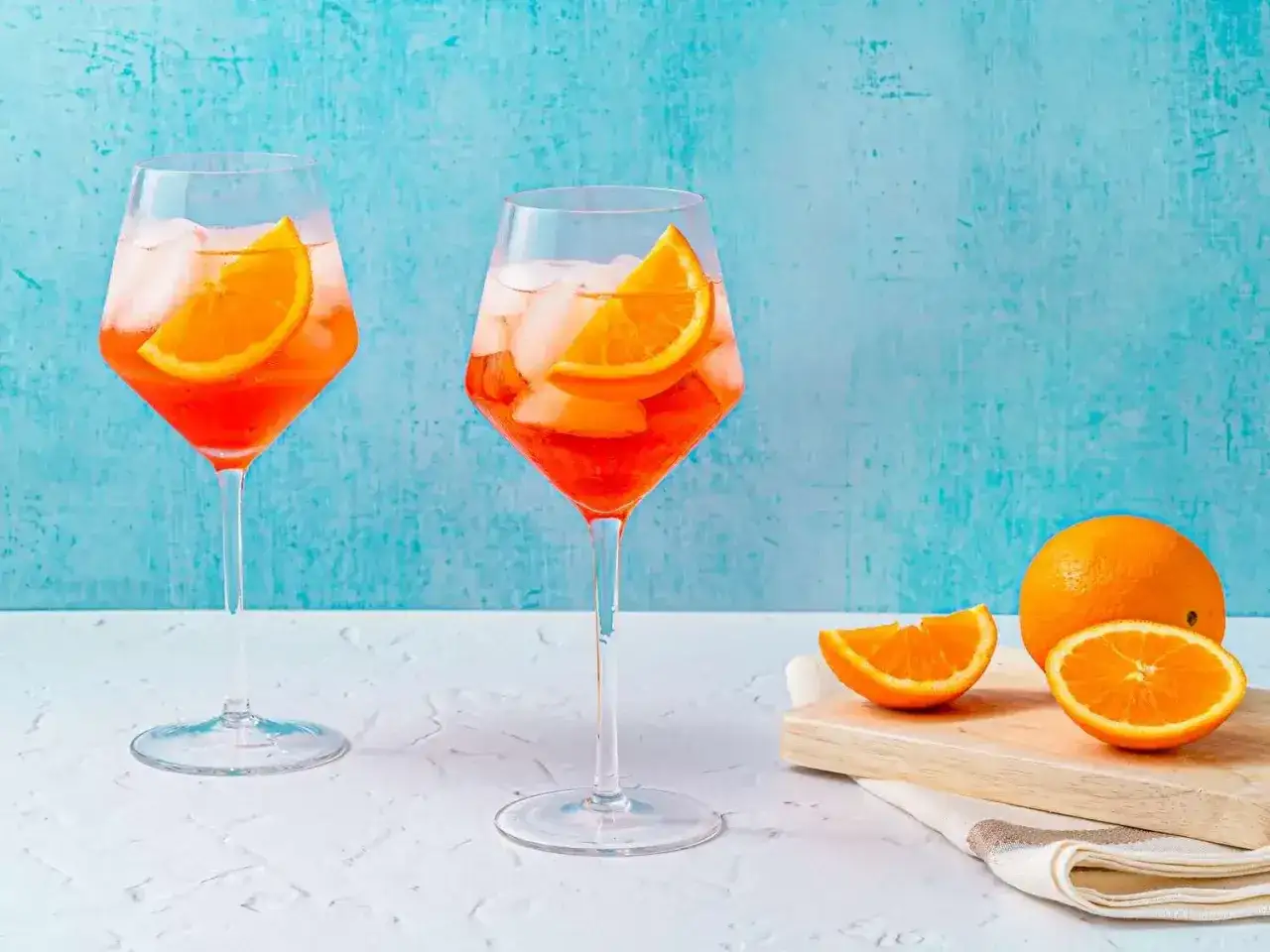 Tv&aring; glas med Aperol Spritz, is och apelsinskivor, med en hel och skivad apelsin p&aring; en sk&auml;rbr&auml;da i bakgrunden.