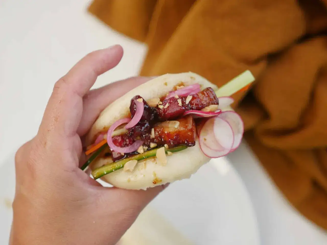 En hand h&aring;ller en bao bun fylld med saftigt fl&auml;sk, krispig gurka, r&ouml;dl&ouml;k och jordn&ouml;tter. Ett perfekt bao buns recept!