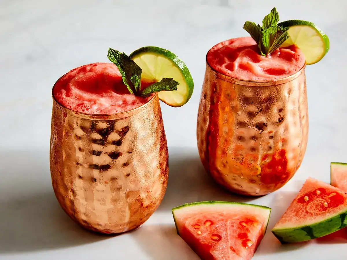 Två frostiga koppar med rosa vodka drinkar, garnerade med lime och mynta. Vattenmelonbitar ligger bredvid.