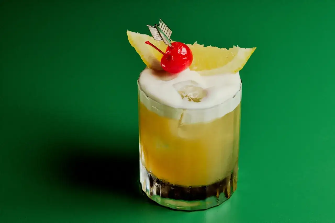 En Amaretto Sour med skummig topp, citronklyfta, k&ouml;rsb&auml;r och en pil.