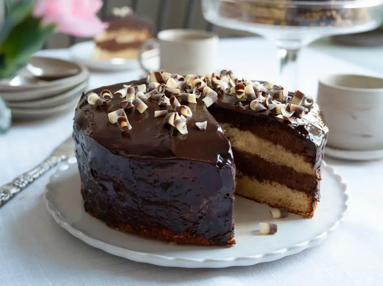 En läcker chokladtårta med en glansig chokladglaze till tårta, toppad med chokladlockar. En bit är uppskuren och visar lager av ljus och mörk choklad.