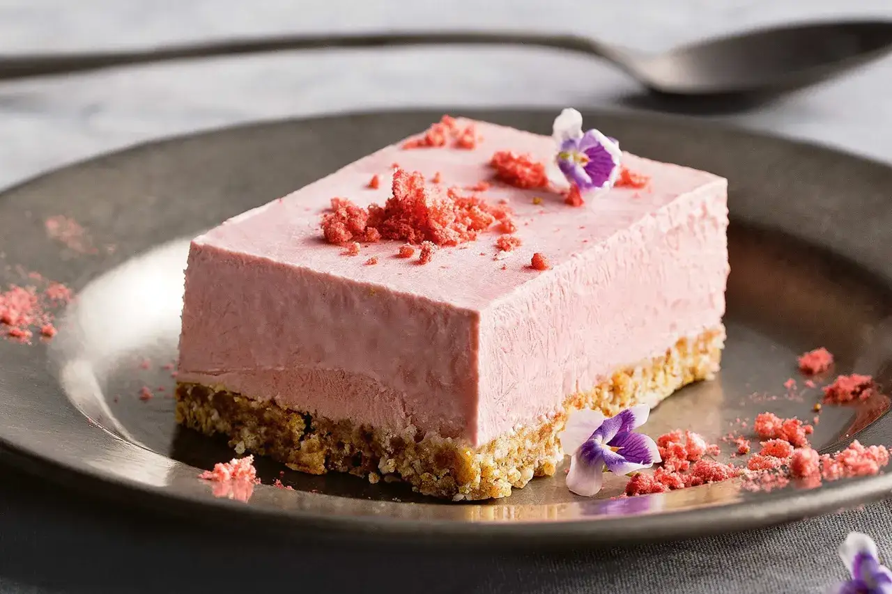 En bit fryst cheesecake med jordgubbssmak, toppad med smulor och blommor.