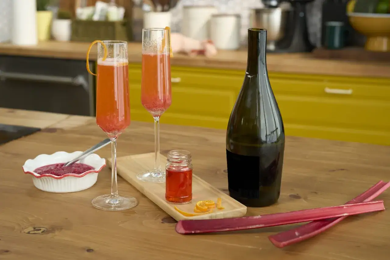 Tv&aring; glas med rabarberdrink och prosecco, en flaska, rabarber och en liten flaska med r&ouml;d v&auml;tska.