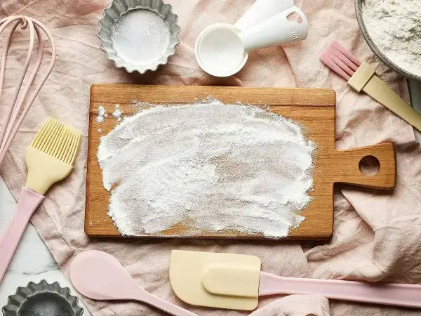 Bakredskap som visp, slickepott och måttskedar ligger utspridda. Ett bakbord har mjöl på sig, redo för att byta ut bikarbonat mot bakpulver.