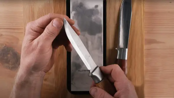Händer som håller en kniv över en slipsten, visar hur slipar man knivar för att få en vass egg. En annan kniv ligger bredvid.